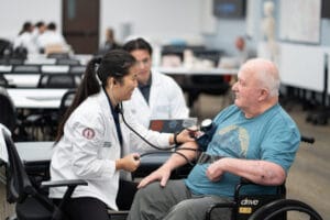 A medical professional checks the blood pressure of an older man in a wheelchair while another medical worker observes in a clinical setting.