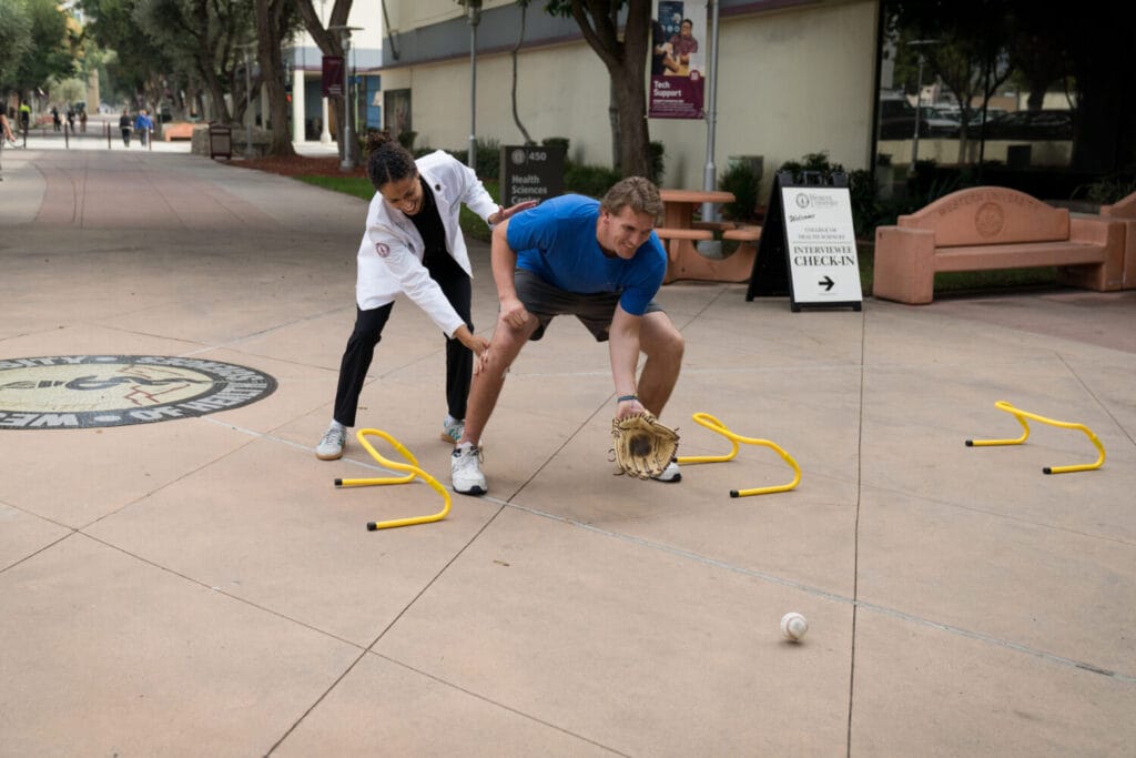 A coach helps a man in athletic clothes field a baseball with a glove while navigating hurdles on a campus sidewalk.