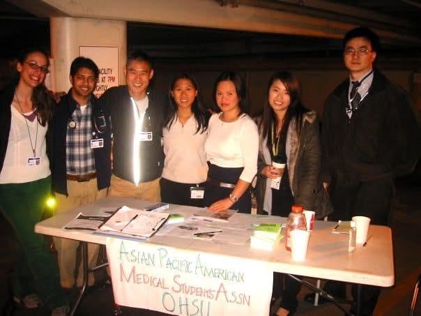 Seven people stand behind a table with papers, cups, and a Gatorade bottle. A banner reads "Asian Pacific American Medical Students Assn OHSU." The group poses and smiles for the photo.