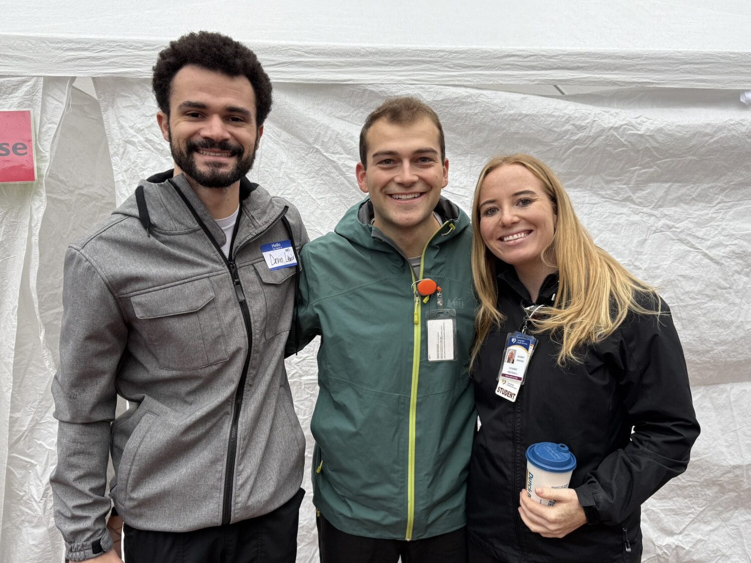 Three people stand side by side smiling in front of a white tent, each wearing jackets and name badges. One person holds a coffee cup.