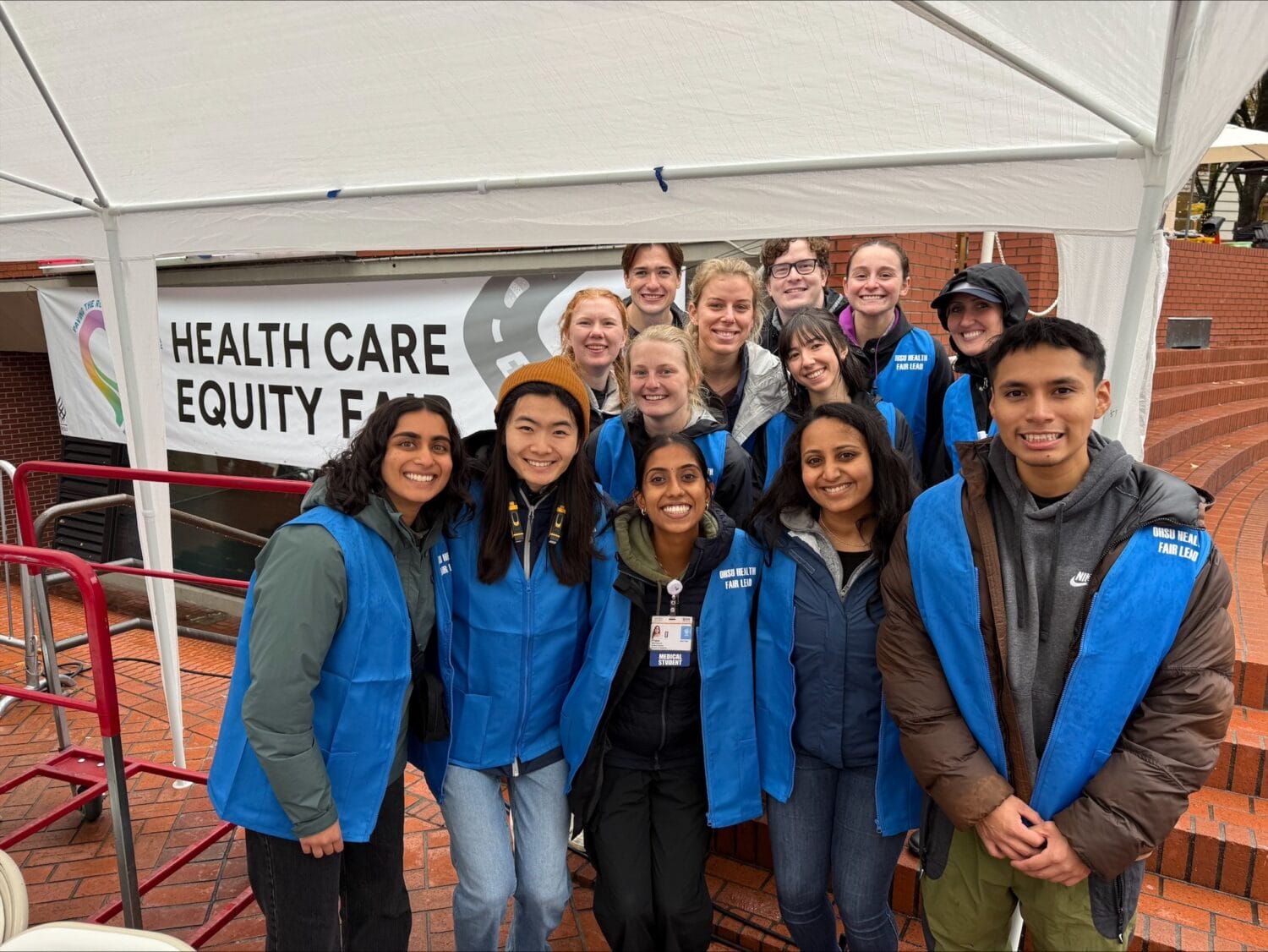 A group of people wearing blue vests pose and smile under a canopy in front of a 