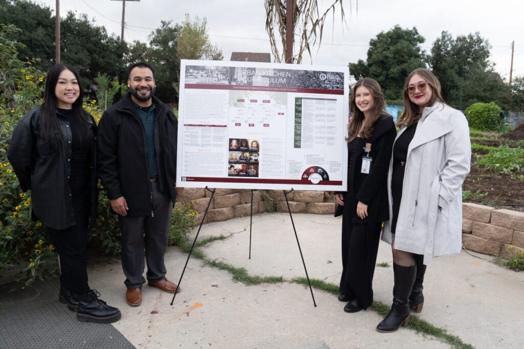 Four people stand outdoors next to a display board with project information and images, with greenery and a stone wall in the background.