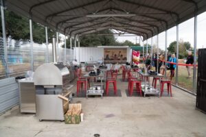 Outdoor covered kitchen setup with stainless steel appliances, red chairs, and several tables; people are gathered near a red ribbon in the background.