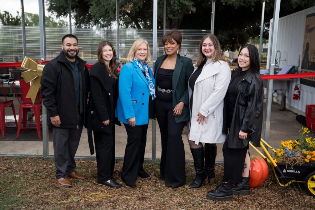 Six people stand side by side outdoors, smiling at the camera. There are trees, red chairs, and a wheelbarrow with flowers in the background.