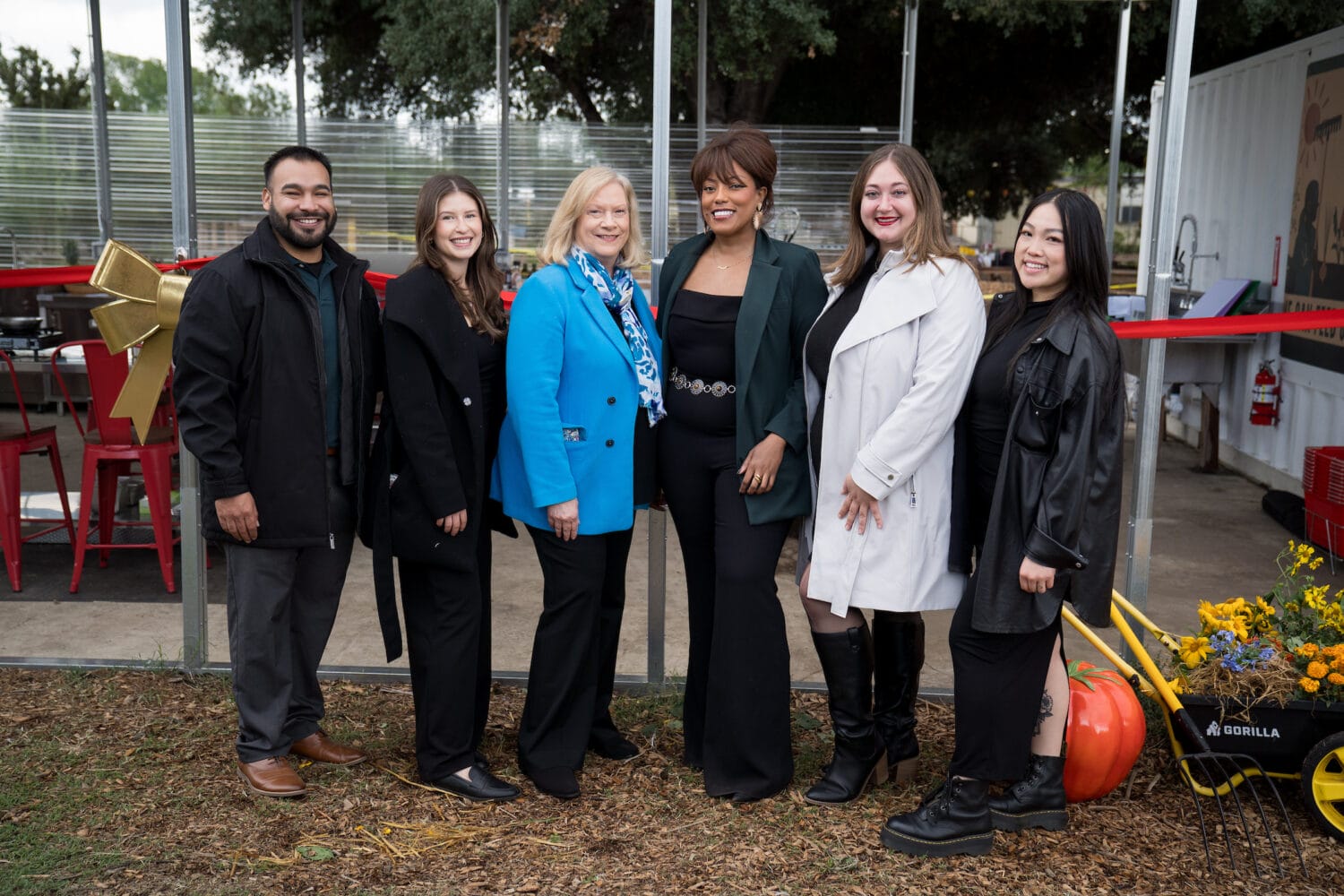 Six people stand side by side outdoors, smiling at the camera. There are trees, red chairs, and a wheelbarrow with flowers in the background.