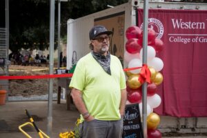 A man in a yellow shirt and bandana stands near a balloon decoration and a red ribbon at an outdoor event with a "Western" university banner in the background.
