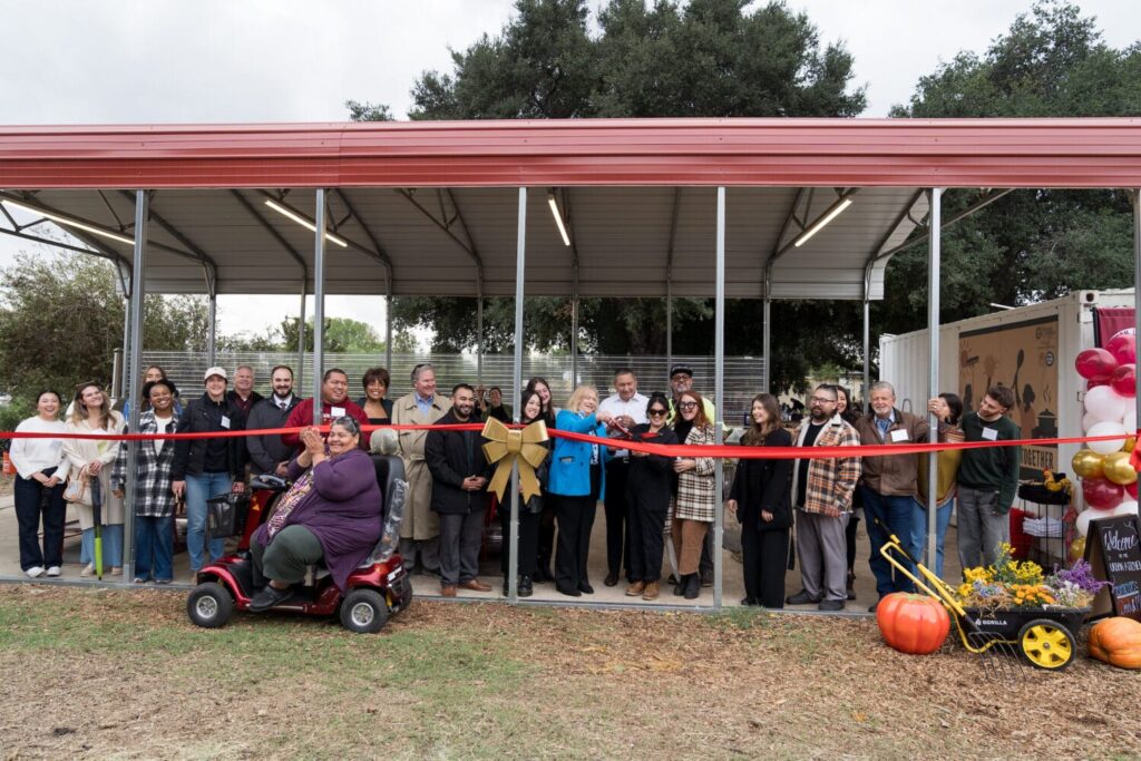 A group of people stand in front of a new open-sided building as a woman in blue cuts a red ribbon at a dedication ceremony. Decorations include autumn produce and balloons.