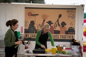 Two women in aprons prepare food at an outdoor cooking station in front of a banner that reads, "We can grow ourselves together.