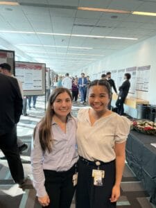 Two women stand side by side and smile at a professional conference or poster session, with other attendees and research posters visible in the background.