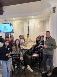 A group of people sit and stand around a table with food and drinks in a restaurant, with a Western University screen visible in the background.