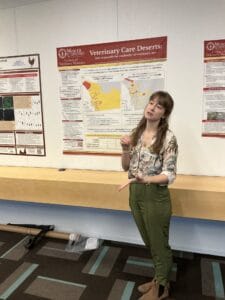 A woman stands in front of a research poster titled "Veterinary Care Deserts" at an academic event, explaining its content.