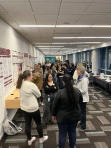 People gather in a hallway for a poster presentation event, viewing research posters on the wall and engaging in discussion. Tables with refreshments are set up along the windows.
