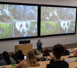 A woman gives a lecture in a classroom with students seated, while a presentation featuring images of animals and text is displayed on two large screens.
