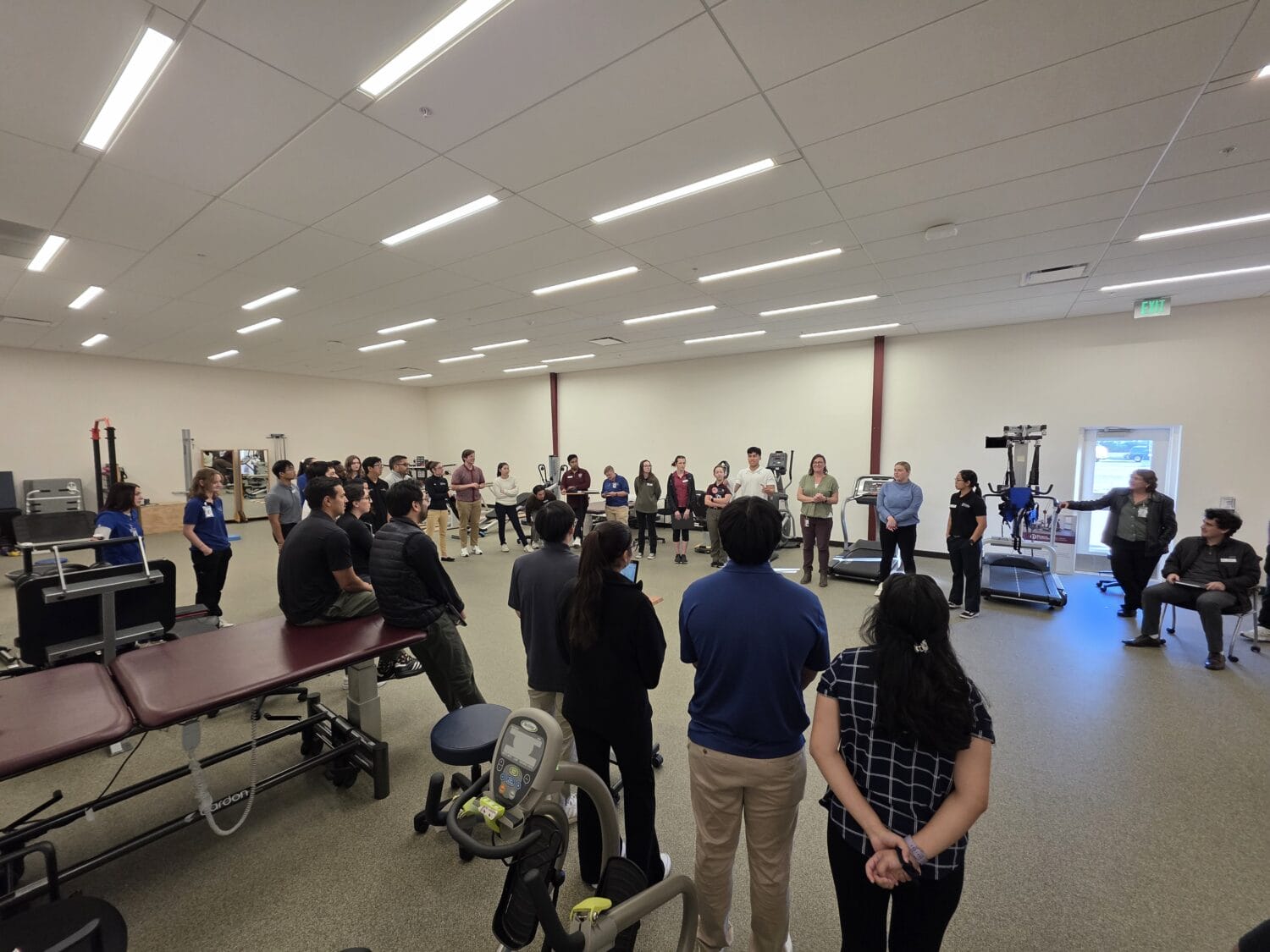 A group of people stands in a large, well-lit room with exercise equipment, gathered in a loose circle, listening to a speaker.