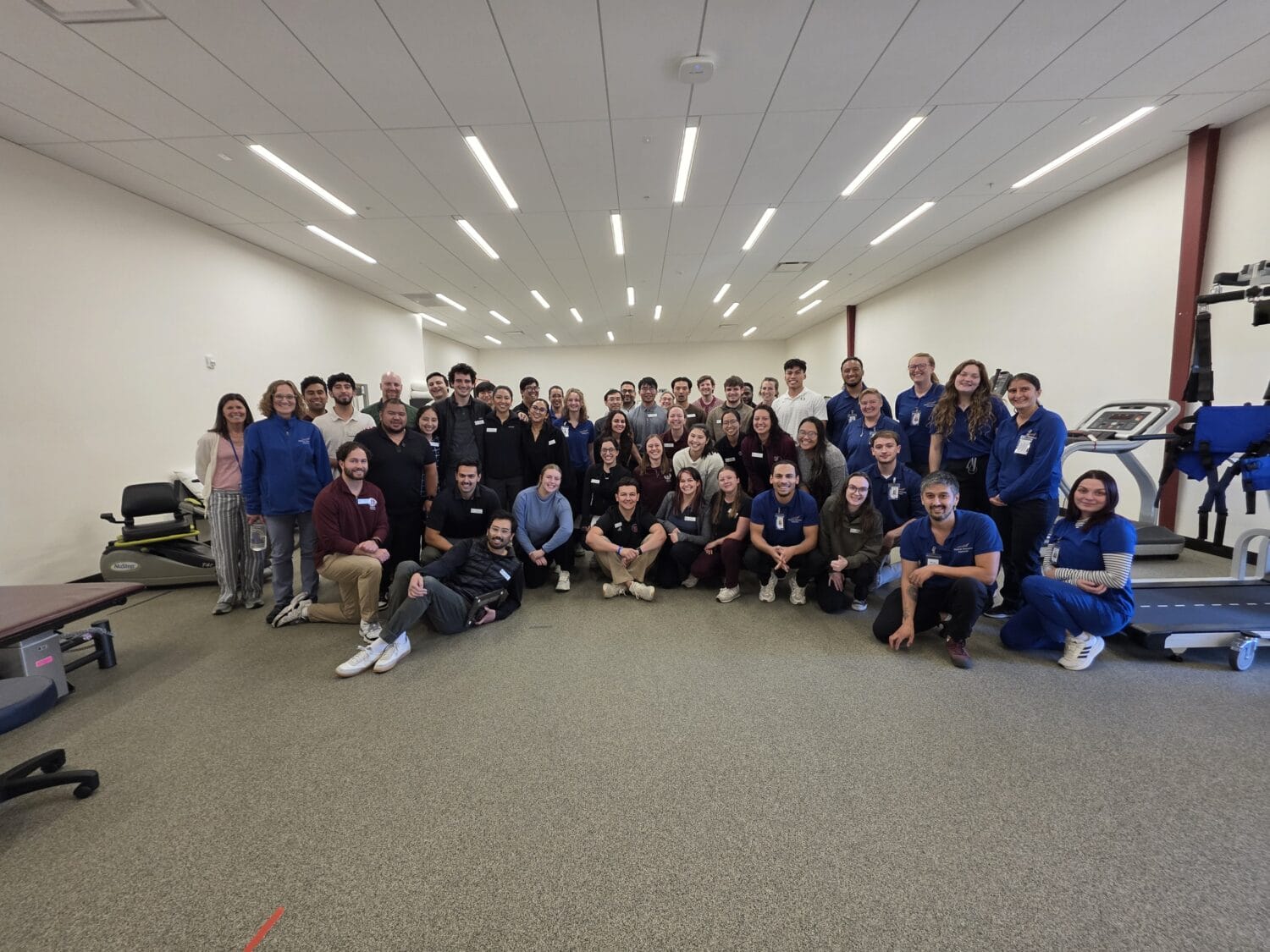 A large group of people pose together in a spacious, well-lit indoor room with medical equipment and exercise machines visible in the background.