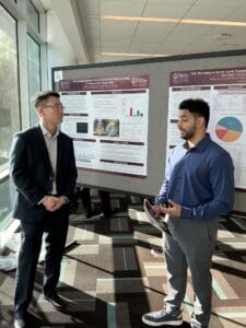 Two men stand and talk in front of scientific research posters at a conference or academic event, with sunlight streaming through large windows.