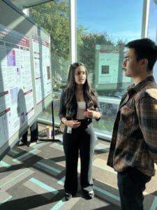 A woman discusses a research poster with a man at an indoor conference or academic event, with natural light coming through large windows.