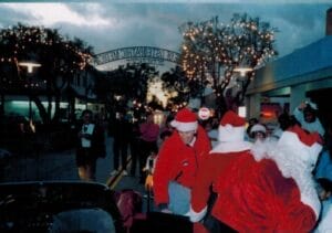 Two people in Santa costumes greet a crowd on a decorated street at dusk; holiday lights and people in festive attire are visible.
