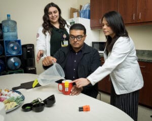 Two women in lab coats assist a seated man as he pours water from a pitcher into a mug on a table in a break room setting.