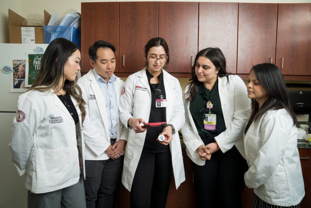 Five healthcare professionals in white coats stand together in an office, looking at a device held by the person in the center.