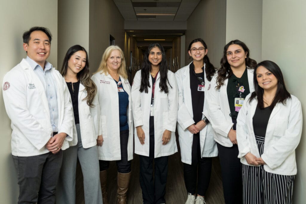 Seven medical professionals in white lab coats stand in a hallway, facing the camera and smiling.