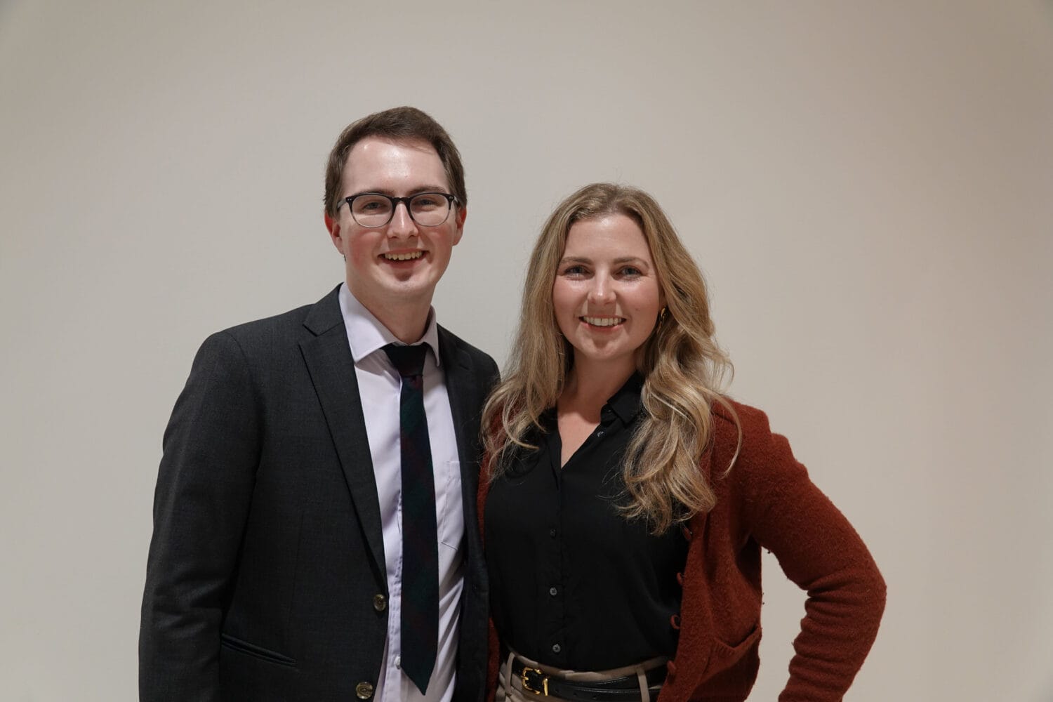 Two people stand side by side, smiling at the camera. The person on the left wears a suit and tie; the person on the right wears a black blouse and red cardigan. Plain background.