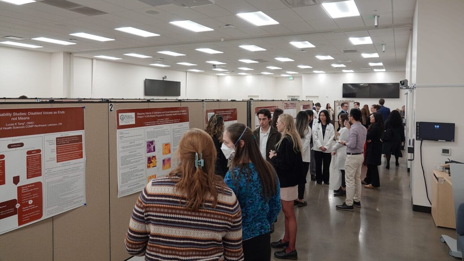 People stand in a well-lit room viewing and discussing research posters displayed on partition walls during a poster presentation event.