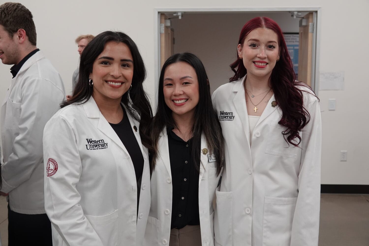 Three women wearing white lab coats stand together and smile inside a building, with two people visible in the background.