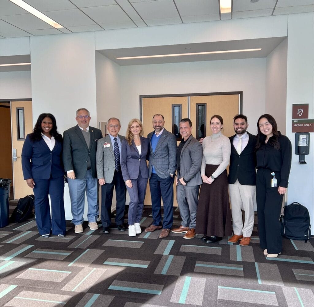 Nine people dressed in business attire stand side by side in a conference room with large windows and patterned carpet, posing for a group photo.