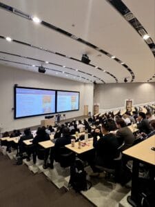 A large lecture hall with students seated and taking notes while a presentation on state authority is displayed on dual screens at the front.