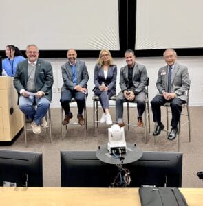 Five people in business attire sit on stools in front of a projection screen, posing for a group photo at what appears to be a panel event or presentation.