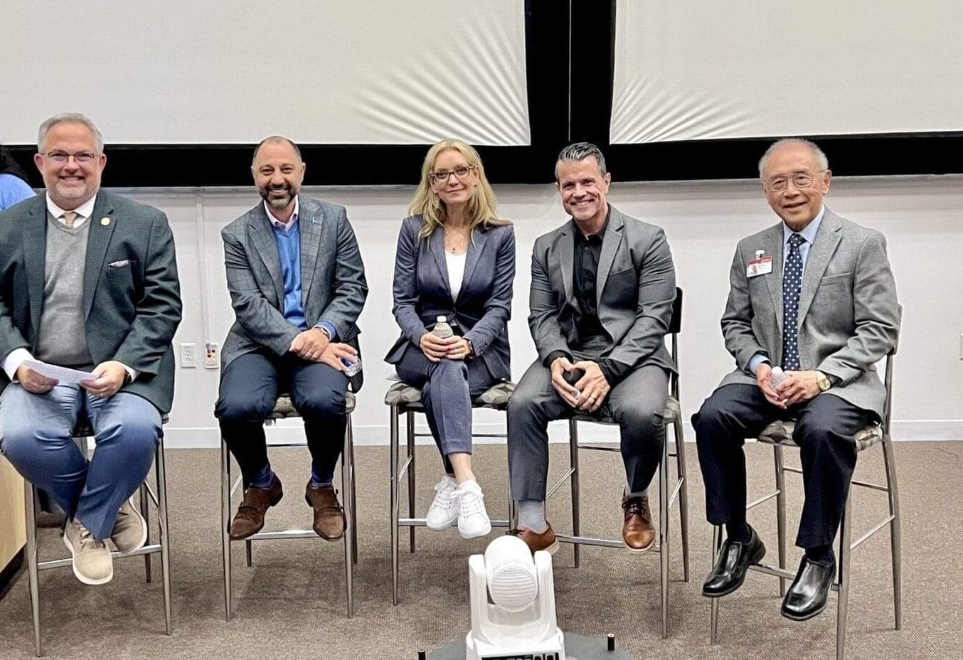 Five people in business attire sit on stools in front of a projection screen, posing for a group photo at what appears to be a panel event or presentation.