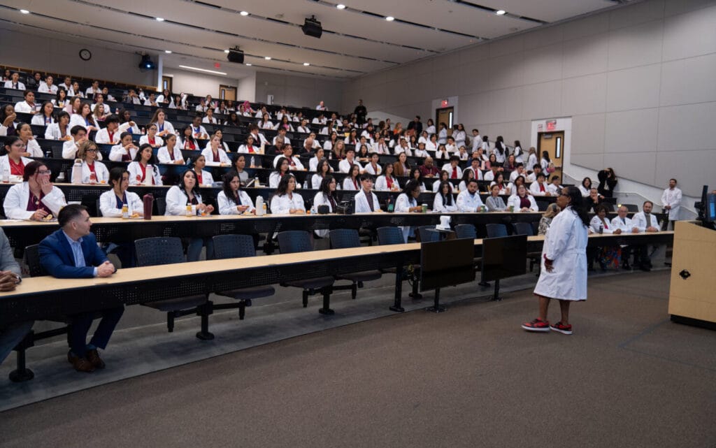 A person in a white coat speaks at the front of a large lecture hall filled with people, most wearing white coats, seated and listening attentively.