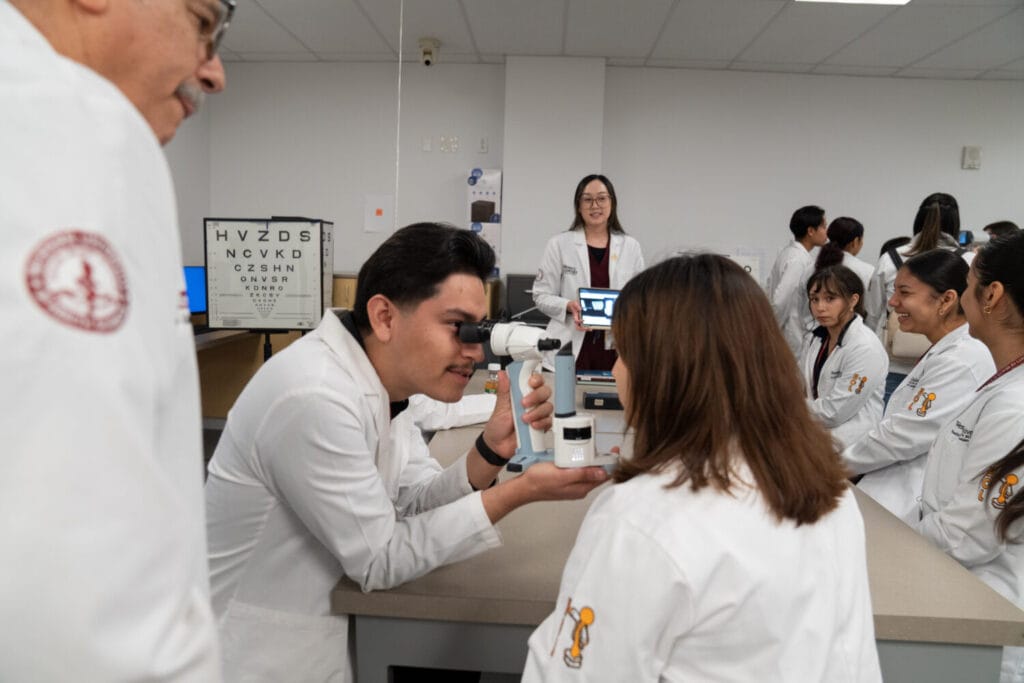 A group of people in white lab coats participate in an eye examination demonstration in a classroom with an eye chart in the background.