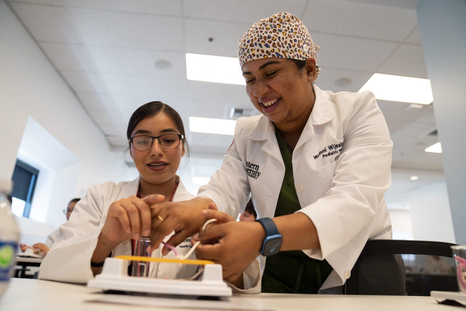 Two women in lab coats practice a medical procedure using a training model at a table in a classroom setting.