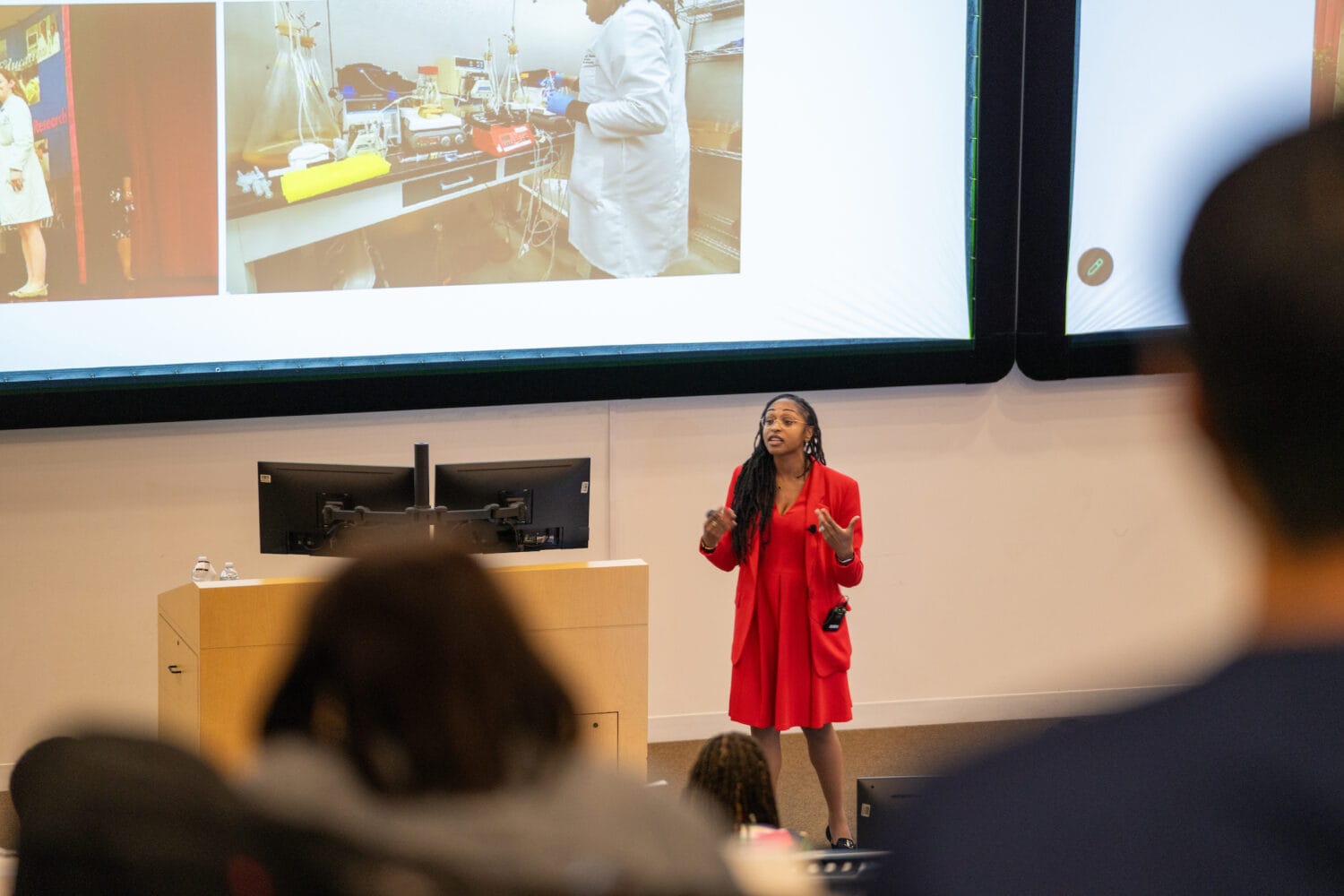 A woman in a red outfit gives a presentation in a lecture hall, with scientific images displayed on a large screen behind her.