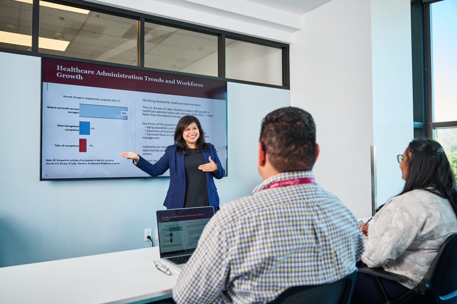 A woman presents healthcare administration trends on a screen to two seated people in a modern classroom setting.