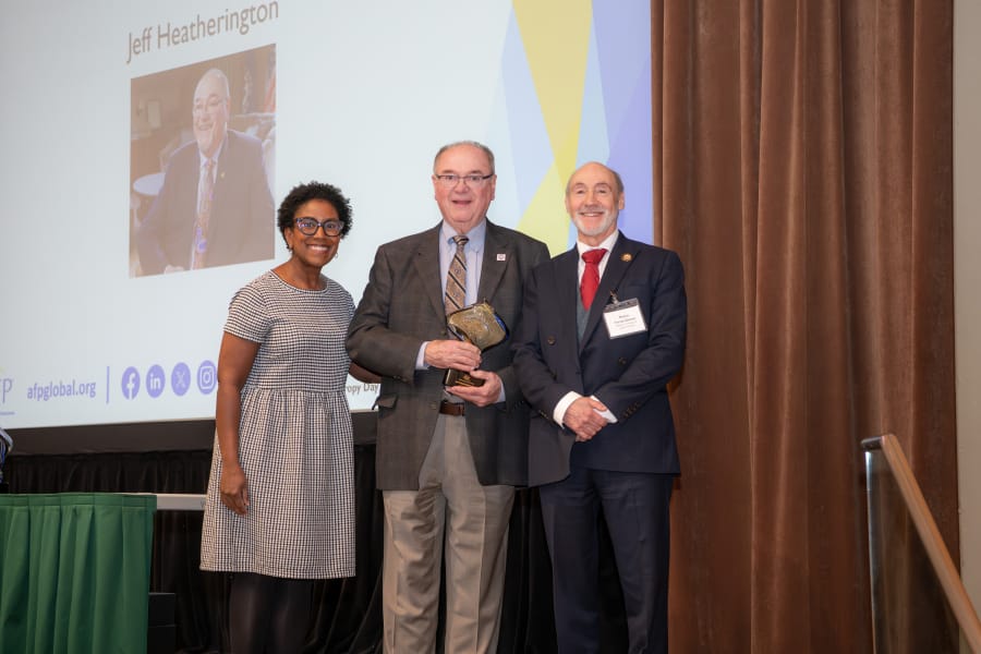 Three people stand together on stage; the man in the center holds an award. A projected slide behind them displays the name 