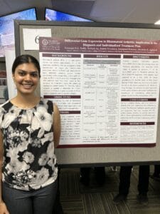 A woman stands beside a scientific poster presentation about differential gene expression in rheumatoid arthritis at a conference or academic event.