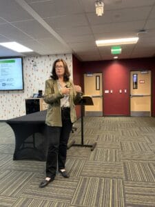 A person stands and speaks in a conference room, gesturing with one hand. A screen with a presentation and a covered table are visible behind them.