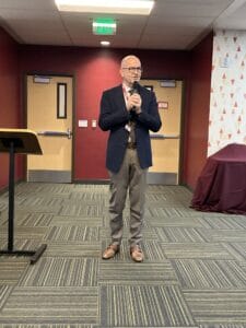 A man in a suit jacket and slacks stands holding a microphone in a conference room with maroon walls and patterned carpet.