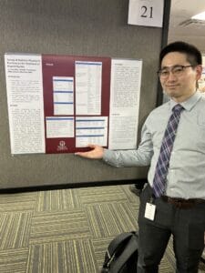 A man wearing glasses and business attire stands indoors, gesturing toward a research poster displayed on a partition.