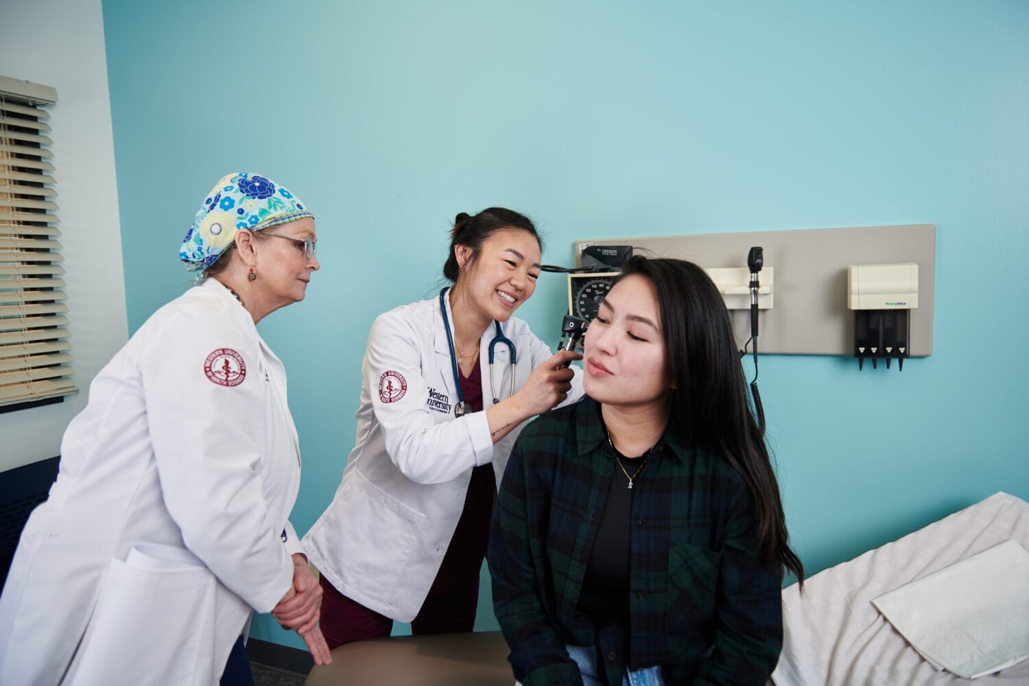 Two healthcare professionals examine a woman's ear in a medical office. The woman is seated on the exam table while one professional uses an otoscope and the other observes.