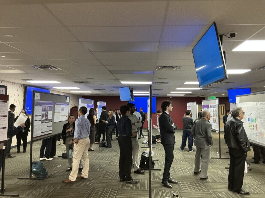 People stand and discuss research posters displayed on boards in a conference room with overhead screens and fluorescent lighting.