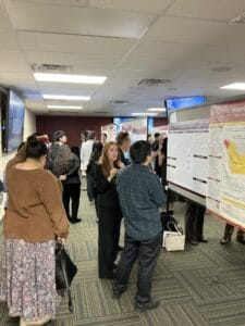 People gather in a room, observing and discussing academic research posters displayed on boards during a poster presentation event.