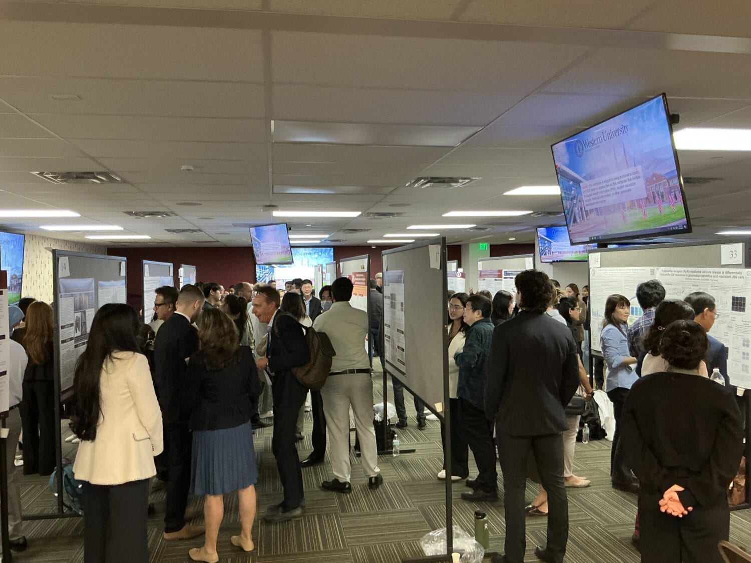 A large group of people in business attire are viewing and discussing research posters in a crowded conference or exhibition hall.