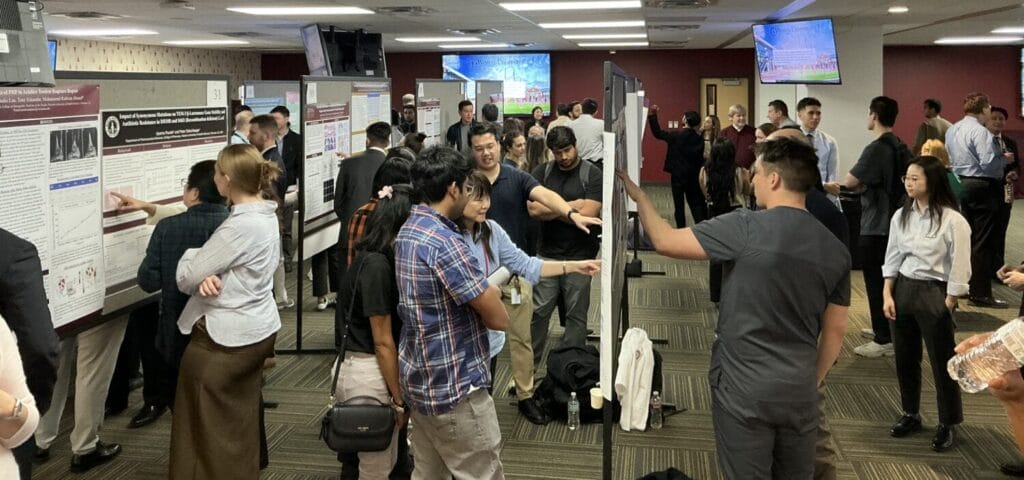 A group of people stand in a room with multiple research posters displayed on boards, engaging in discussions during a poster presentation event.