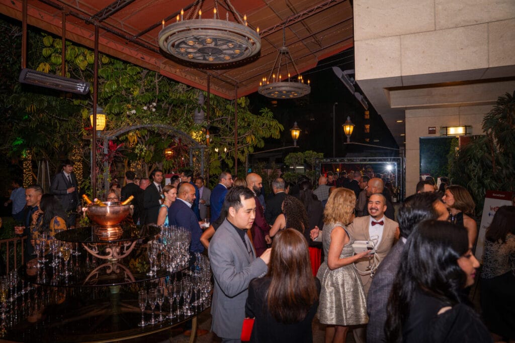 A large group of people in formal attire socialize at an outdoor evening event with greenery, chandeliers, and a table of champagne glasses.