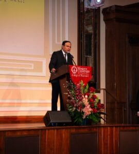 A man in a suit speaks at a podium with a Western University College of Pharmacy sign, while another man stands to the side, holding a folder.