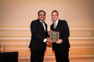 Two men in suits stand side by side, smiling, as one hands the other a plaque at an indoor awards event.